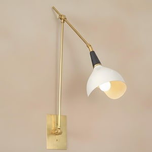 Op de afbeelding: Een wandlamp met een messing afwerking en een witte, klokvormige kap. De lamp heeft een zwart accent en een verstelbare arm. Het licht is aan en verlicht de binnenkant van de kap.