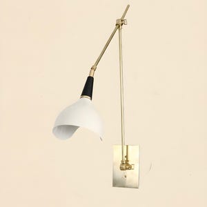 Op de afbeelding: Een wandlamp in mid-century modern stijl, met een witte, gebogen kap en een zwart-gouden arm. De lamp heeft een rechthoekige basis in messingkleur en een verstelbare arm. Het ontwerp is minimalistisch en stijlvol.