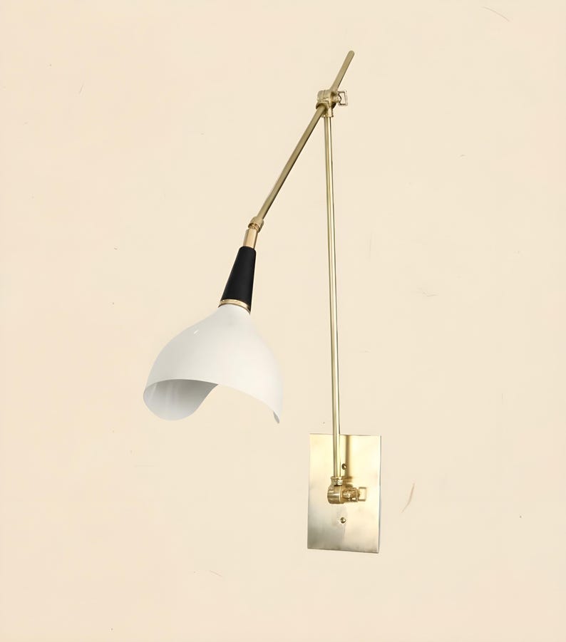 Op de afbeelding: Een wandlamp in mid-century modern stijl, met een witte, gebogen kap en een zwart-gouden arm. De lamp heeft een rechthoekige basis in messingkleur en een verstelbare arm. Het ontwerp is minimalistisch en stijlvol.