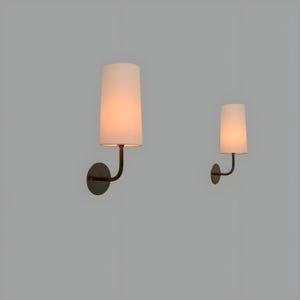 May include: Two wall sconces with white tapered shades and dark metal arms and circular backplates. The sconces are lit, emitting a warm glow. The design is minimalist and modern, suitable for interior lighting.