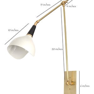 Op de afbeelding: Een moderne wandlamp met een witte kap, zwarte en gouden accenten en een messing afwerking. De lamp strekt zich uit over 38 cm, met een kap van 25 cm. De basis is 10 x 15 cm en de arm is 51 cm lang.