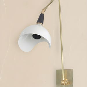 Op de afbeelding: Een moderne wandlamp met een witte, gebogen kap en een zwart-gouden arm. De lamp heeft een messingkleurige rechthoekige basis en een lange, rechte arm. De kap is aan de onderkant open en onthult de gloeilamp.