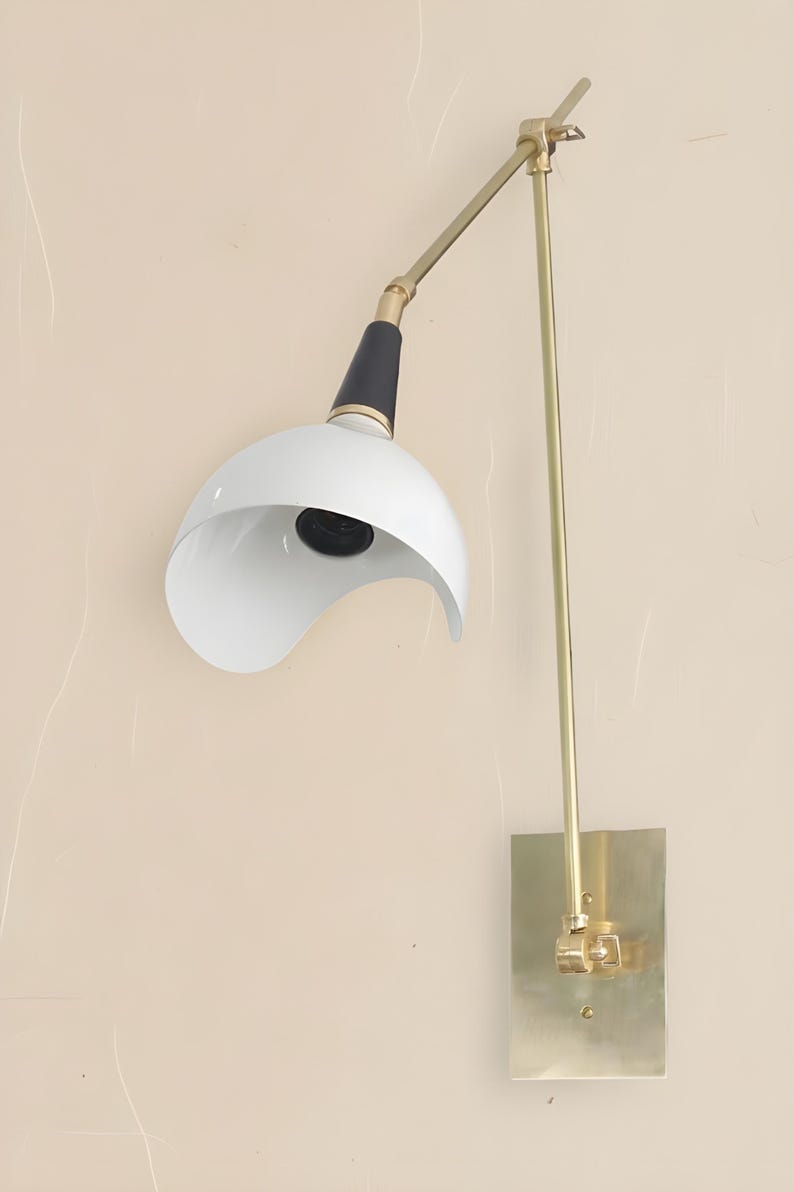 Op de afbeelding: Een moderne wandlamp met een witte, gebogen kap en een zwart-gouden arm. De lamp heeft een messingkleurige rechthoekige basis en een lange, rechte arm. De kap is aan de onderkant open en onthult de gloeilamp.