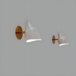 Könnte beinhalten: Zwei weiße Wandleuchten mit messingfarbener Montageplatte und Arm. Die Leuchten haben eine geschwungene, tropfenförmige Form und strahlen ein weiches, nach unten gerichtetes Licht aus. Das Design ist minimalistisch und modern, geeignet für die Innenbeleuchtung.