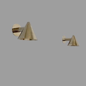 May include: Two brass-coloured wall sconces. Each sconce features a conical shade and a circular mounting plate. The sconces are made of a shiny metal and are mounted on a grey wall. The sconces are different sizes.