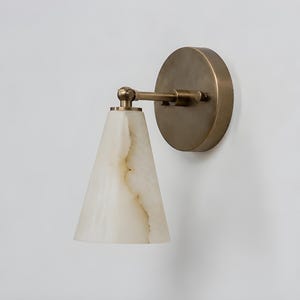 May include: A wall-mounted sconce featuring a conical, off-white stone shade with natural veining. The fixture includes a circular brass backplate and a horizontal arm with an adjustable swivel joint connecting to the stone lampshade.