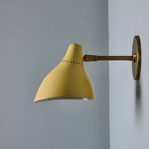 Pode incluir: Uma luminária de parede amarela com uma cúpula curva, braço de latão e base circular. A cúpula tem pequenos orifícios ao longo da borda superior. A luminária está montada em uma parede cinza claro. Dimensões: aproximadamente 30 cm de comprimento.