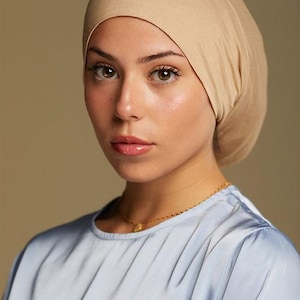 May include: A woman wearing a light beige head covering and a light blue satin top. A delicate gold necklace adorns the neck. The head covering is smooth and form-fitting. The background is a solid, neutral color.
