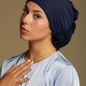 May include: A navy blue head covering with a gathered design. The head covering is worn with a light blue satin top and a gold necklace. The person is wearing gold earrings and has long, manicured nails.