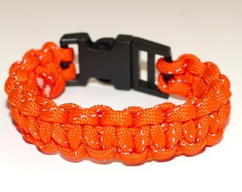 May include: Orange paracord bracelet with a black plastic buckle. The bracelet has reflective threads woven into the paracord.