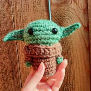 May include: A crocheted figure of a character with large ears, green skin, and black eyes. The figure is wearing a brown robe. The figure is being held by a hand. The background is a wooden surface.