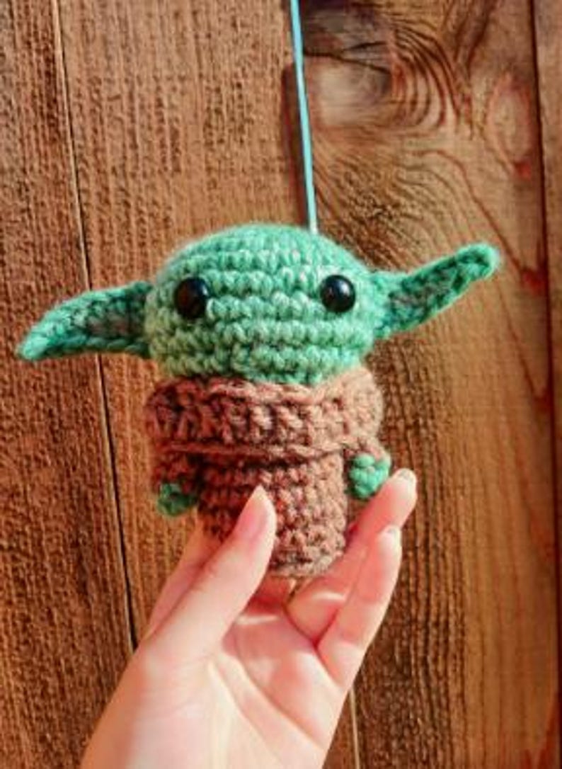 May include: A crocheted figure of a character with large ears, green skin, and black eyes. The figure is wearing a brown robe. The figure is being held by a hand. The background is a wooden surface.