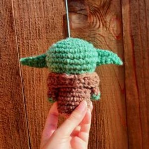 May include: A crocheted figure of a character with a large head and pointed ears, in shades of green and brown. The figure is suspended against a wooden background. The figure is approximately 3 inches tall.