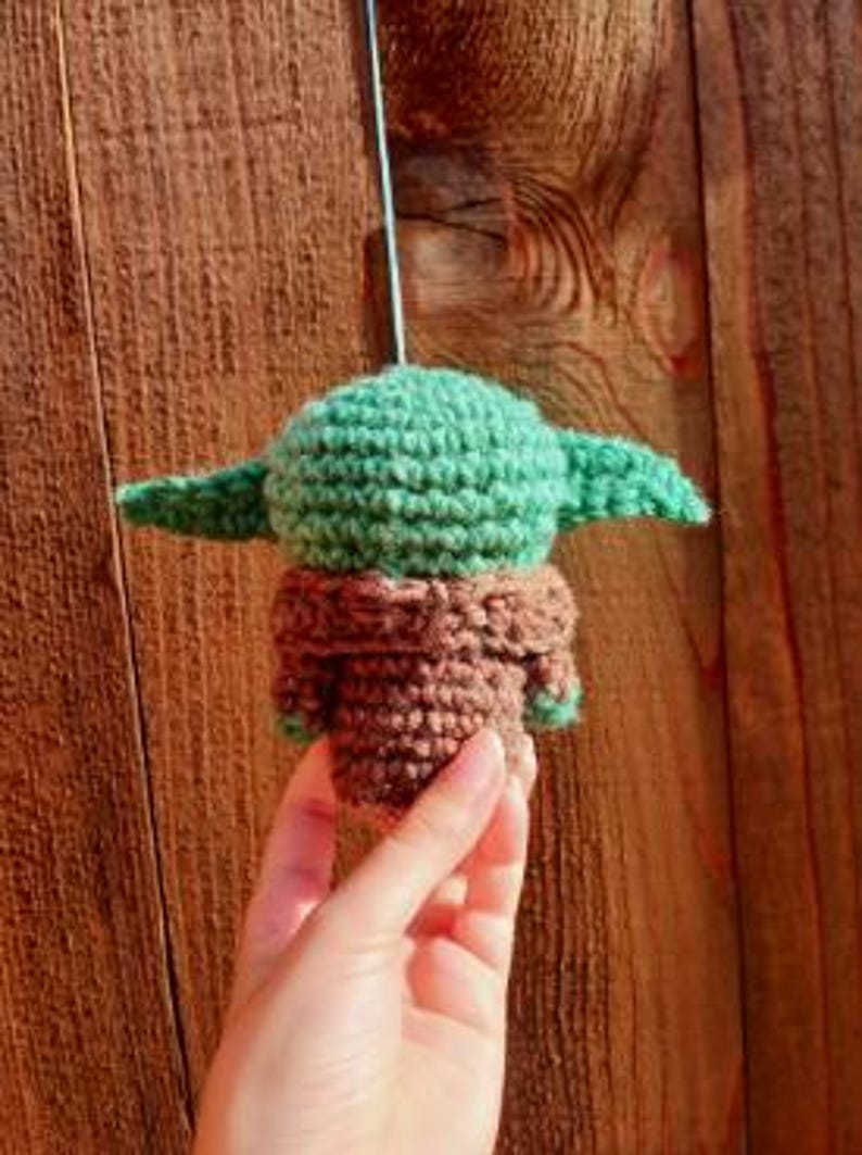May include: A crocheted figure of a character with a large head and pointed ears, in shades of green and brown. The figure is suspended against a wooden background. The figure is approximately 3 inches tall.