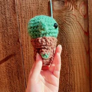 May include: A small, crocheted green creature with a brown base, held against a wooden background. The creature has a round head, black eyes, and a small heart-shaped detail on the base. The crochet work is visible, showing the texture of the yarn.