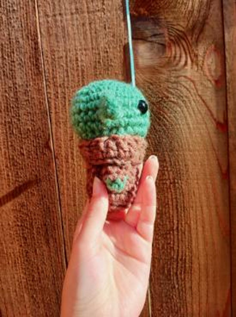 May include: A small, crocheted green creature with a brown base, held against a wooden background. The creature has a round head, black eyes, and a small heart-shaped detail on the base. The crochet work is visible, showing the texture of the yarn.