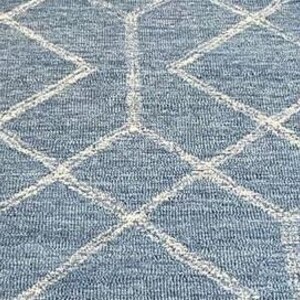 Hand tufted wool area rug with a prominent white geometric diamond pattern on a textured slate blue background. The handmade carpet is laid flat on a dark hardwood floor in a luxury living room, partially styled under a cream-colored sectional sofa and glass coffee table, capturing the complete pattern and premium texture.