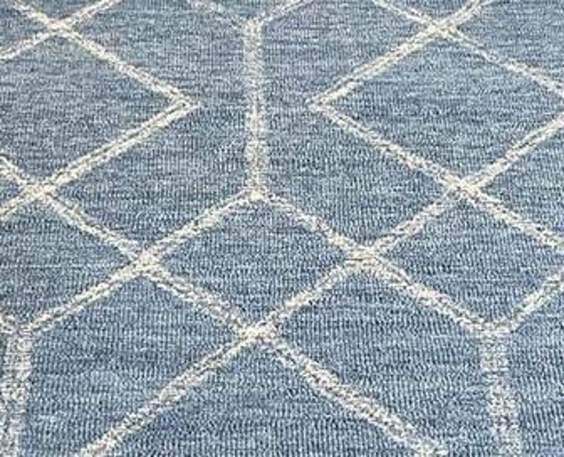 Hand tufted wool area rug with a prominent white geometric diamond pattern on a textured slate blue background. The handmade carpet is laid flat on a dark hardwood floor in a luxury living room, partially styled under a cream-colored sectional sofa and glass coffee table, capturing the complete pattern and premium texture.