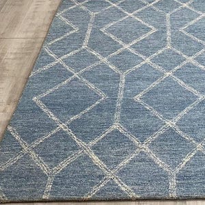 Hand tufted wool area rug with a prominent white geometric diamond pattern on a textured slate blue background. The handmade carpet is laid flat on a dark hardwood floor in a luxury living room, partially styled under a cream-colored sectional sofa and glass coffee table, capturing the complete pattern and premium texture.