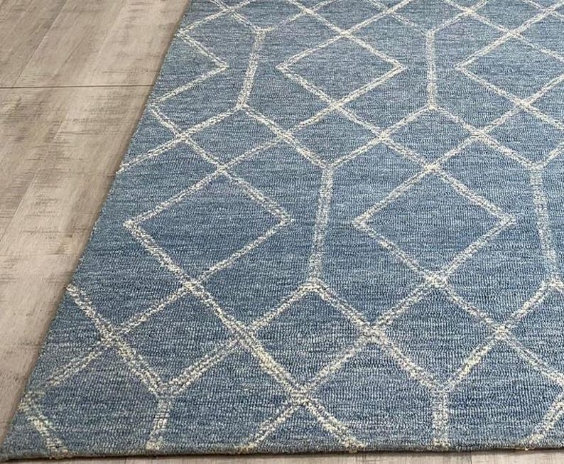 Hand tufted wool area rug with a prominent white geometric diamond pattern on a textured slate blue background. The handmade carpet is laid flat on a dark hardwood floor in a luxury living room, partially styled under a cream-colored sectional sofa and glass coffee table, capturing the complete pattern and premium texture.