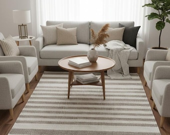 Puede incluir: Salón con un sofá gris claro, cojines de tonos neutros, dos sillones a juego y una mesa de centro redonda de madera. Una alfombra a rayas grises y blancas cubre el suelo. Una lámpara y una planta en maceta completan la decoración.