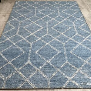 Hand tufted wool area rug with a prominent white geometric diamond pattern on a textured slate blue background. The handmade carpet is laid flat on a dark hardwood floor in a luxury living room, partially styled under a cream-colored sectional sofa and glass coffee table, capturing the complete pattern and premium texture.