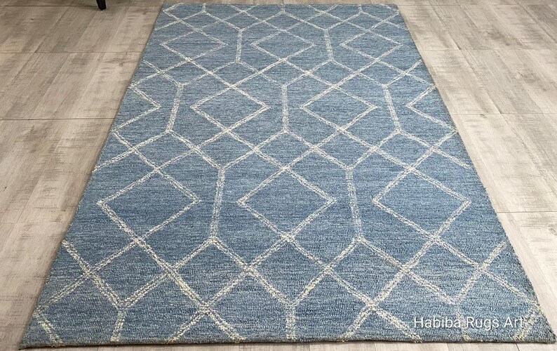 Hand tufted wool area rug with a prominent white geometric diamond pattern on a textured slate blue background. The handmade carpet is laid flat on a dark hardwood floor in a luxury living room, partially styled under a cream-colored sectional sofa and glass coffee table, capturing the complete pattern and premium texture.