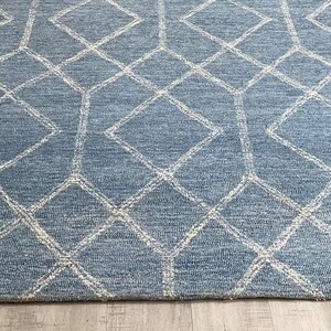 Hand tufted wool area rug with a prominent white geometric diamond pattern on a textured slate blue background. The handmade carpet is laid flat on a dark hardwood floor in a luxury living room, partially styled under a cream-colored sectional sofa and glass coffee table, capturing the complete pattern and premium texture.