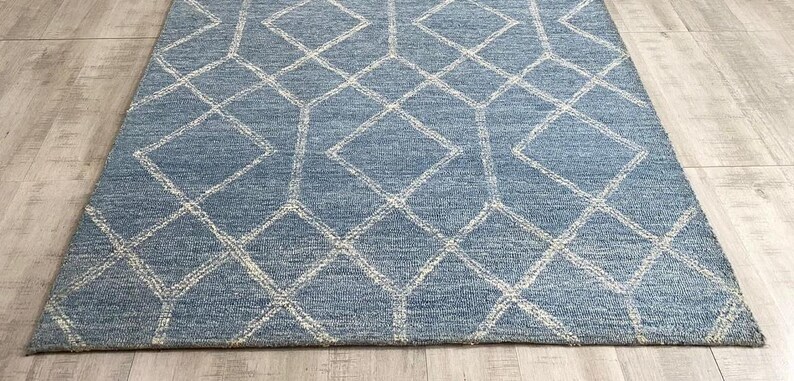 Hand tufted wool area rug with a prominent white geometric diamond pattern on a textured slate blue background. The handmade carpet is laid flat on a dark hardwood floor in a luxury living room, partially styled under a cream-colored sectional sofa and glass coffee table, capturing the complete pattern and premium texture.