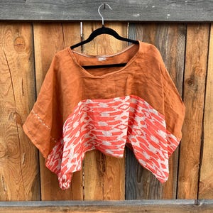May include: An orange linen top with a wide neckline and short sleeves. The bottom section features a white and coral abstract pattern. The top is hanging on a black hanger against a wooden background.