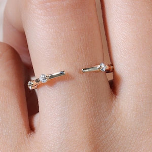 May include: A gold-colored open ring with small, clear gemstones. The ring has a split design, with the gemstones set along the band. The ring is shown on a person's finger.