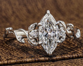 May include: A silver-toned ring featuring a large marquise-cut center stone held by prongs. The band is designed with intricate, raised leaf-like metalwork patterns that extend along the sides, accented by two small round stones on each side.