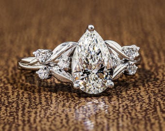 May include: A silver-toned ring featuring a large pear-shaped center stone held by prongs. The band is designed with metallic leaf-like motifs that wrap around the center stone, accented by several small, round, clear stones set along the sides.