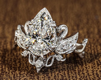 May include: A silver-toned ring featuring a large pear-cut center stone surrounded by smaller round-cut stones. The metal band is crafted with intricate, curved, and vine-like detailing that frames the central pear-shaped gemstone.