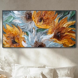 May include: An oil painting of sunflowers in shades of yellow, orange, white, and blue. The artwork is displayed in a black frame above a bed with white linens. The painting's textured brushstrokes create a three-dimensional effect.