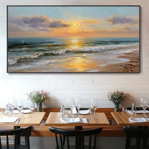 Puede incluir: Pintura al óleo enmarcada que representa una puesta de sol sobre el océano. El cielo transita del azul al naranja, con nubes. Las olas rompen en la playa arenosa, reflejando la luz dorada. Debajo, mesas de restaurante con cristalería y arreglos florales.
