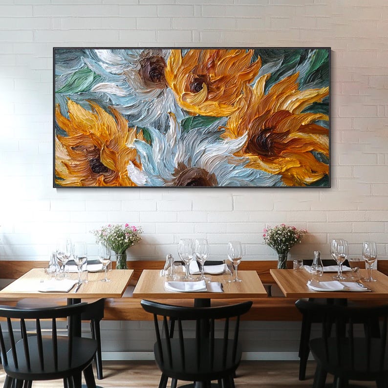 May include: A framed oil painting of sunflowers in shades of yellow, orange, white, and blue. The artwork is displayed above a restaurant table setting with chairs, glassware, and floral arrangements. The painting's textured brushstrokes are visible.