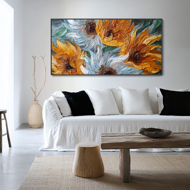 May include: An oil painting of sunflowers in shades of yellow, orange, and white, set against a backdrop of blue and green. The artwork is framed in black and hangs above a white sofa with black and white pillows. A wooden coffee table and a beige rug complete the scene.