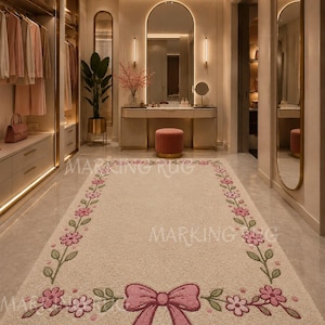 Puede incluir: Una alfombra rectangular de color crema con un borde de flores rosas con tallos y hojas verdes. Un lazo rosa grande y texturizado está centrado en el borde inferior de la alfombra. El texto MARKING RUG es visible en una fuente clara.