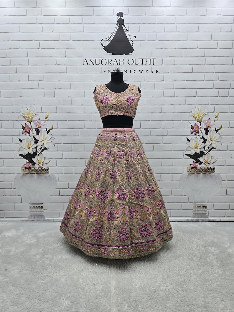 May include: A two-piece formal outfit on a mannequin. The outfit features a sleeveless, pink embroidered top and a matching full skirt. The skirt and top are adorned with intricate floral patterns in shades of pink, gold, and purple. The brand name "ANUGRAH OUTFIT" is visible.