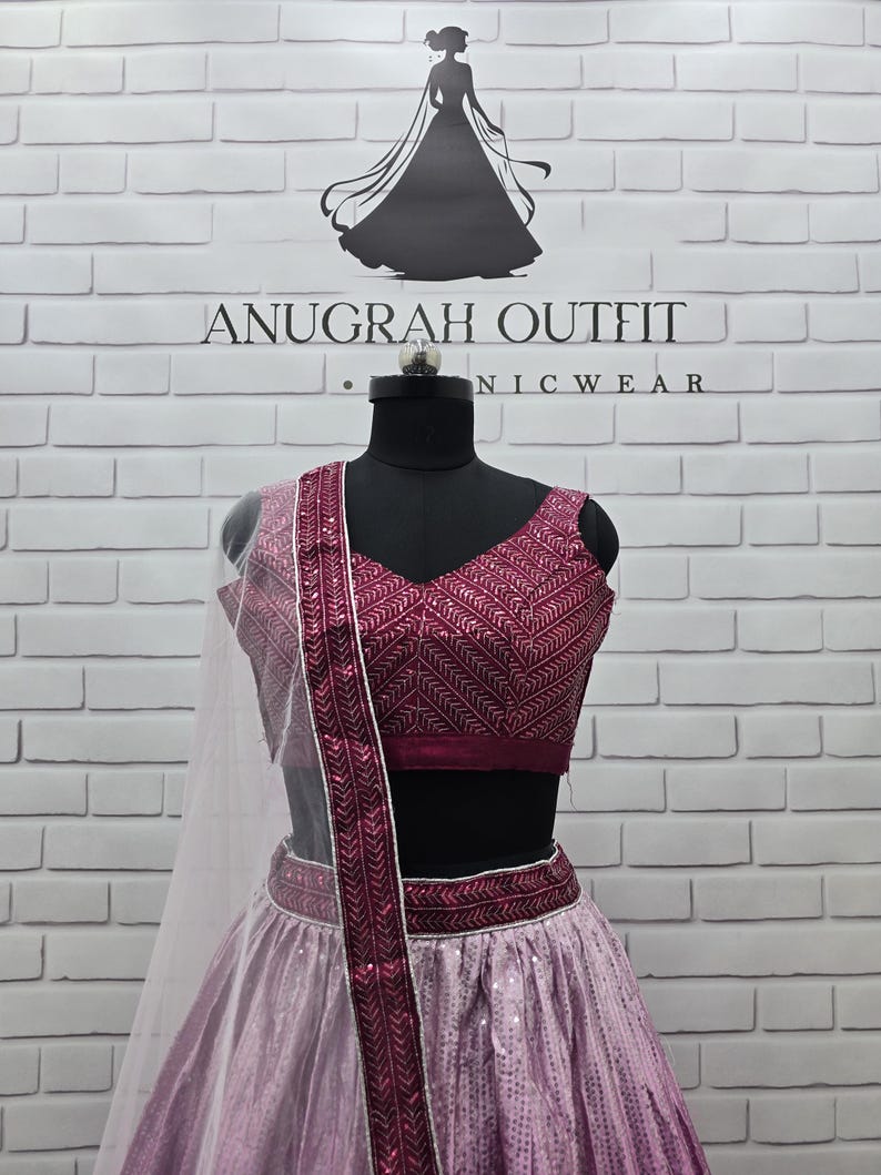 May include: A formal, two-piece outfit on a mannequin. The top is a deep magenta with intricate embroidery, paired with a matching skirt. The skirt has a sequined design. A sheer, white scarf is draped over the shoulder. The text "ANUGRAH OUTFIT NICWEAR" is visible.