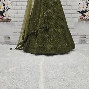 May include: Olive green formal gown with intricate embroidery and a matching sheer shawl. The gown features a detailed pattern across the skirt and bodice, with subtle embellishments. The fabric drapes elegantly, creating a flowing silhouette.