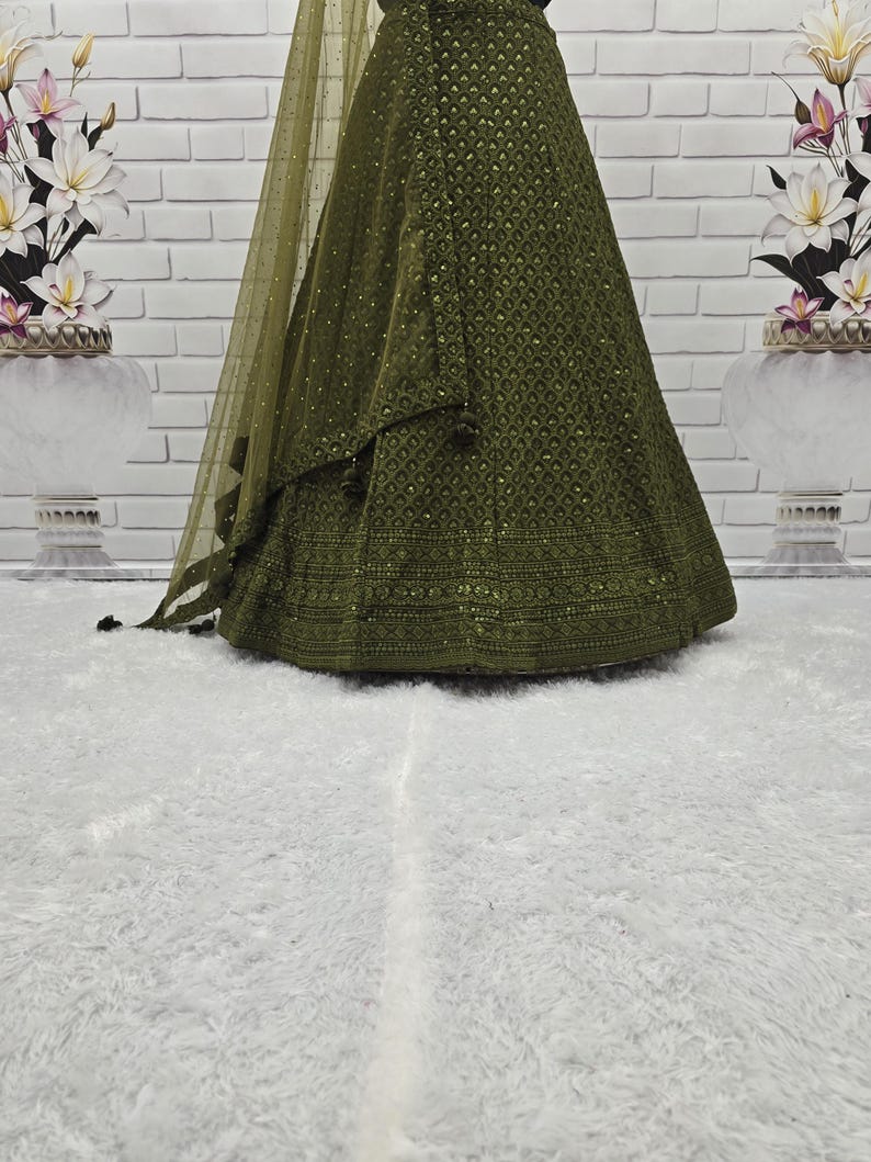 May include: Olive green formal gown with intricate embroidery and a matching sheer shawl. The gown features a detailed pattern across the skirt and bodice, with subtle embellishments. The fabric drapes elegantly, creating a flowing silhouette.