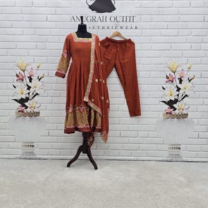 May include: A rust-colored three-piece outfit consisting of a long-sleeved, knee-length dress with gold embroidery at the neckline, cuffs, and hem, matching trousers, and a coordinating dupatta with gold accents, displayed on a mannequin.