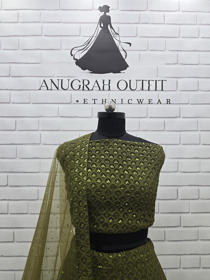 May include: Olive green ethnic wear ensemble on a mannequin. The outfit features intricate sequin detailing and a sheer, matching dupatta. The background includes the text "ANUGRAH OUTFIT &bull; ETHNICWEAR" and a silhouette of a woman.
