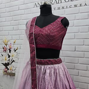 May include: A formal outfit on a mannequin. The ensemble features a deep burgundy top with intricate embroidery, a matching waistband, and a flowing skirt that fades from light pink to a deeper shade. A sheer, white scarf with burgundy trim completes the look. The text "ANURAH OUTFIT" is visible.