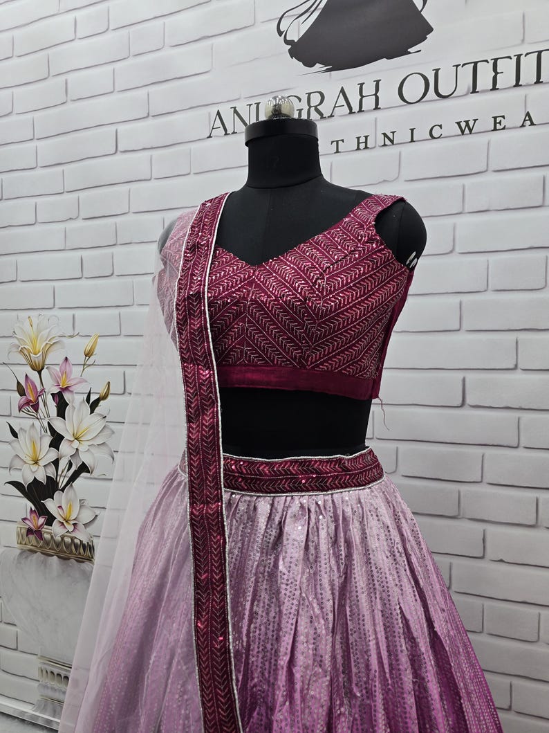 May include: A formal outfit on a mannequin. The ensemble features a deep burgundy top with intricate embroidery, a matching waistband, and a flowing skirt that fades from light pink to a deeper shade. A sheer, white scarf with burgundy trim completes the look. The text "ANURAH OUTFIT" is visible.