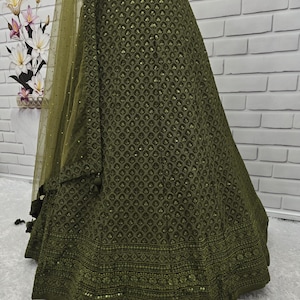 May include: Olive green skirt with intricate embroidered patterns and a matching sheer dupatta. The skirt features a detailed border design and a flowing silhouette. The fabric has a subtle sheen, and the overall look is elegant and sophisticated.
