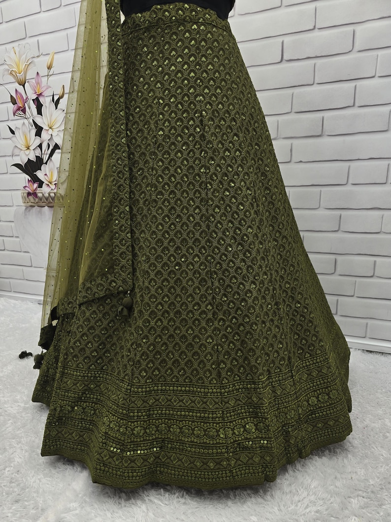 May include: Olive green skirt with intricate embroidered patterns and a matching sheer dupatta. The skirt features a detailed border design and a flowing silhouette. The fabric has a subtle sheen, and the overall look is elegant and sophisticated.