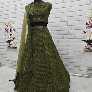 May include: An olive green ethnic outfit, including a crop top and a long skirt, displayed on a mannequin. The outfit features intricate patterns and a matching sheer scarf. The background is a white brick wall. The text "ANUGRAH OUTFIT" is visible at the top.
